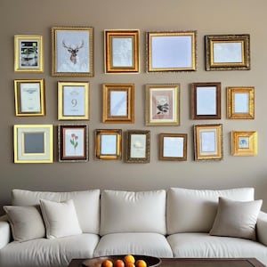 May include: A display of gold-framed pictures and artwork of various sizes and shapes, arranged on a neutral-coloured wall. The frames have ornate details and are hung above a white sofa with neutral-coloured cushions.