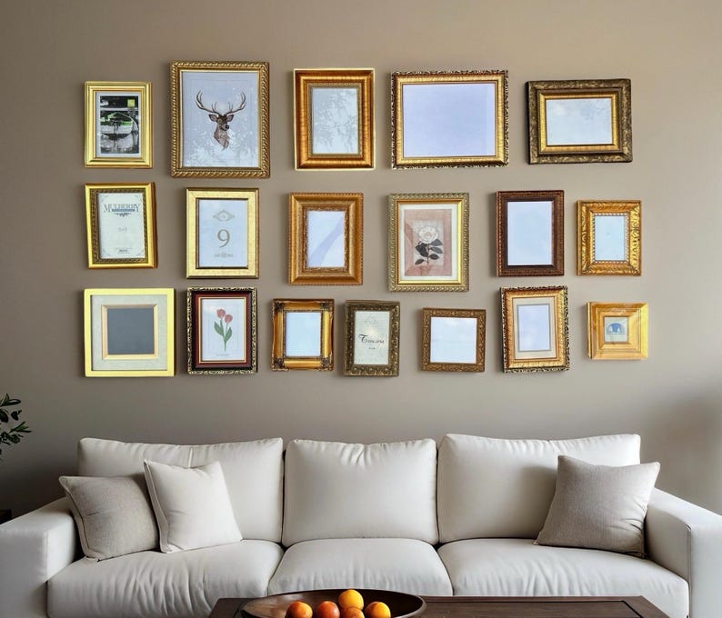 May include: A display of gold-framed pictures and artwork of various sizes and shapes, arranged on a neutral-coloured wall. The frames have ornate details and are hung above a white sofa with neutral-coloured cushions.