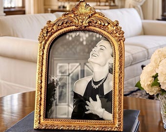 May include: A gold-colored ornate picture frame with a black and white photo of a woman. The frame is resting on a dark book titled "ANNE OF GREEN GABLES". The frame has an arched top and is decorated with intricate details.