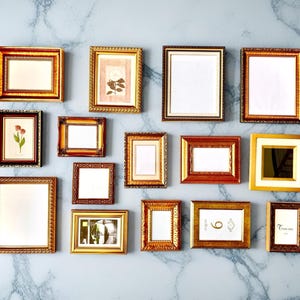 May include: A collection of various gold and brown picture frames of different sizes and shapes, arranged on a marble-patterned wall. Some frames contain artwork, while others are empty.