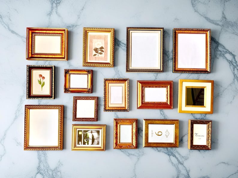 May include: A collection of various gold and brown picture frames of different sizes and shapes, arranged on a marble-patterned wall. Some frames contain artwork, while others are empty.