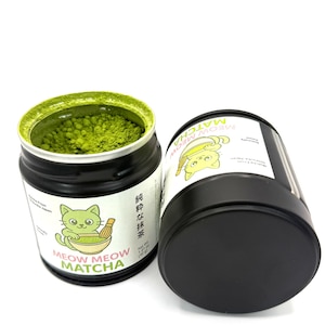May include: Two black metal tins of matcha powder. One tin is open, revealing bright green powder. The tins have labels with a cartoon cat and the text "MEOW MEOW MATCHA". The open tin is filled with matcha powder, and the other tin is lying on its side.