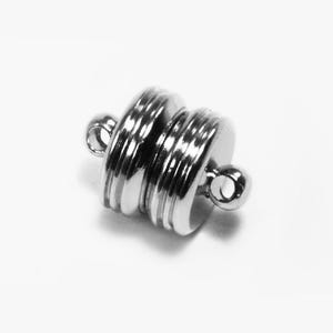 Può includere: Una chiusura magnetica argentata con un design cilindrico. La chiusura presenta una serie di scanalature orizzontali e piccoli anelli su ciascuna estremità. La chiusura è progettata per collegare due pezzi.