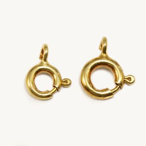Könnte beinhalten: Zwei goldfarbene Federringverschlüsse, essentielle Komponenten für die Schmuckherstellung. Jeder Verschluss hat ein kreisförmiges Design mit einem kleinen Hebel zum Öffnen und Schließen sowie eine Schlaufe zum Befestigen.