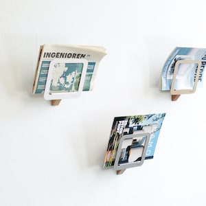 Könnte beinhalten: Drei Wand-Zeitschriftenhalter. Jeder Halter hat eine Holzbasis mit einem weißen Metallrahmen, der eine Zeitschrift oder Zeitung hält. Die Zeitschriften haben farbige Umschläge und Text. Die Halter sind an einer weißen Wand befestigt.