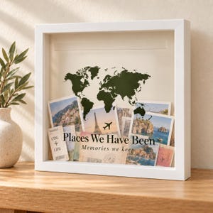 May include: A white square shadow box frame containing a dark green world map silhouette and various travel photographs, boarding passes, and postcards. The text "Places We Have Been" and "Memories we keep" is printed on the glass front.