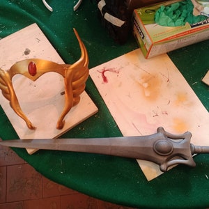 Puede incluir: Una corona dorada con una gema roja y una espada gris se exhiben sobre una superficie de madera. La corona tiene detalles en forma de alas. La espada tiene una empuñadura detallada y una hoja larga. Accesorios de cosplay.