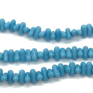 May include: Three strands of light blue, elongated beads are displayed against a white background. Each bead has a rounded shape, and they are strung together. The beads are attached to a silver tag.