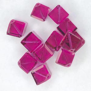 May include: A collection of faceted, square-shaped beads in a vibrant magenta hue. Each bead features a diagonal internal cut, creating a geometric design. The beads have small holes for stringing, ideal for jewelry making or crafting projects.