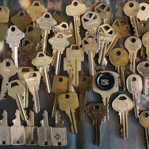 May include: A collection of vintage keys in various styles and finishes, including brass, nickel, and chrome. Some keys have brand names such as Schlage, Yale, and Kwikset. The keys are arranged in a random pattern on a textured surface.