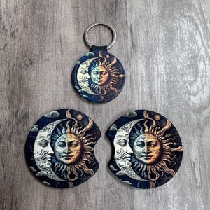May include: A set of three round coasters featuring a sun and moon design. The coasters are black with a blue, white, and gold design. The sun and moon have faces and are surrounded by clouds and stars. The set includes a matching keychain.
