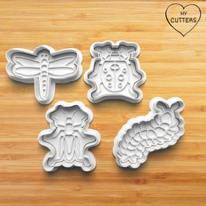 May include: Four white plastic cookie cutters shaped like a dragonfly, ladybird, bow, and caterpillar. The cutters are on a wooden surface. The text "MY CUTTERS" is inside a heart shape in the upper right corner.
