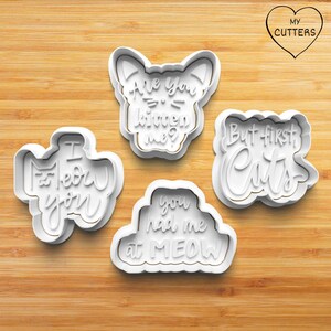 May include: Set of five white plastic cookie cutters with cat-themed sayings. The cutters are shaped like a cat, a heart, and two different banners. The sayings on the cutters are "Are you kitten me?", "I meow you", "You had me at meow", and "But first, cats".