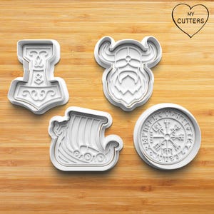 Peut inclure: Ensemble de quatre emporte-pièces en plastique blanc en forme de navire viking, de casque viking, de boussole viking et de marteau viking. Les emporte-pièces sont sur une surface en bois.