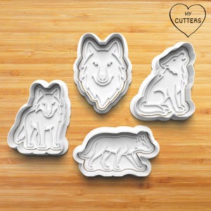 May include: Four white plastic wolf-shaped cookie cutters. The cutters show a wolf's head, a howling wolf, a wolf sitting, and a wolf walking. The cutters are on a wooden surface. The text "MY CUTTERS" is in a heart-shaped logo.