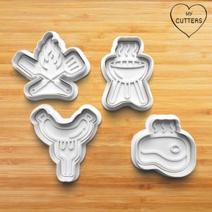 Könnte beinhalten: Vier weiße Ausstecher aus Kunststoff in Form von Grill-Artikeln. Die Formen umfassen ein Lagerfeuer mit gekreuzten Utensilien, einen Grill, eine Wurst mit einer Gabel und ein Steak. Die Ausstecher befinden sich auf einer Holzoberfläche.