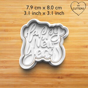 Może przedstawiać: Biała plastikowa foremka do ciastek w kształcie słów Happy New Year napisanych kursywą. Wymiary wynoszą 7,9 cm na 8,0 cm. Tekst MY CUTTERS jest widoczny wewnątrz serca w prawym górnym rogu obrazu.