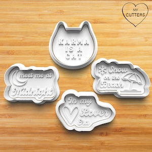 Könnte beinhalten: Vier weiße Ausstecher mit verschiedenen Formen und Texten. Einer ist ein Katzenkopf mit "Karma is a Cat", ein anderer sagt "meet me at Midnight", ein dritter sagt "In my Lover Era" und der letzte sagt "Snow on the Beach".