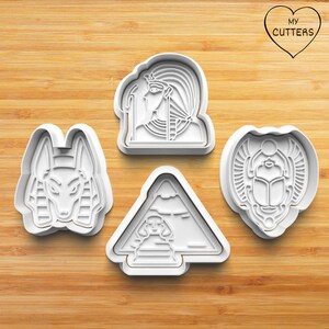 Könnte beinhalten: Ein Set aus vier weißen Kunststoff-Ausstechformen mit ägyptischen Motiven: ein Schakalkopf, ein Pharaonenprofil, eine Pyramide mit Sphinx und ein Skarabäus-Käfer. Jede Form besitzt eine erhöhte Kante zum Ausstechen und geprägte Details.