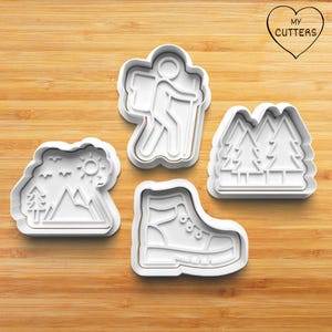 Könnte beinhalten: Vier weiße Ausstecher aus Kunststoff in Outdoor-Abenteuer-Formen: ein Wanderer mit Rucksack, eine Berglandschaft mit Bäumen und Sonne, ein Paar Kiefern und ein Wanderschuh. Die Ausstecher befinden sich auf einer Holzoberfläche.