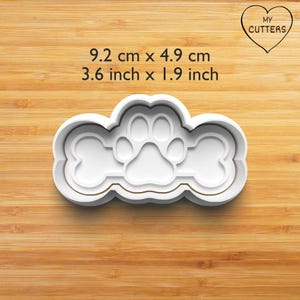 Könnte beinhalten: Ein weißer Ausstechform aus Kunststoff mit einem zentralen Pfotenabdruck, der von zwei Knochenformen flankiert wird. Der Text MY CUTTERS steht in einem Herz. Die Maße betragen 9,2 cm mal 4,9 cm. Die Form hat einen erhöhten Rand zum Ausstechen.