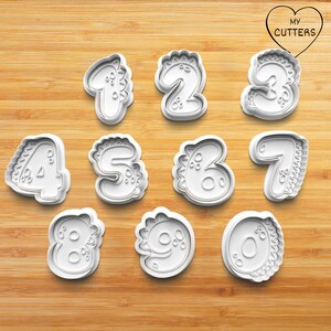 Könnte beinhalten: Ein Satz von zehn weißen Kunststoff-Ausstechern in Form von Zahlen, jeweils mit einem dinosaurierartigen Design. Die Zahlen 1 bis 9 und 0 sind abgebildet. Die Ausstecher liegen auf einer Holzoberfläche. Das Logo "MY CUTTERS" ist herzförmig.