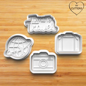 May include: Four white plastic cookie cutters in travel-themed shapes: a globe with a plane, a camera, a suitcase, and the text "Time To Travel". The cutters are on a wooden surface.