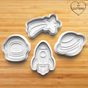 May include: Four white plastic cookie cutters in space-themed shapes: an astronaut helmet, a rocket ship, a shooting star, and a planet with rings. The cutters are on a wooden surface.