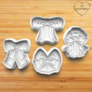 May include: Four white plastic cookie cutters shaped like bows. The cutters are on a wooden surface. The text "MY CUTTERS" is in a heart shape in the top right corner of the image.
