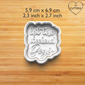 Op de afbeelding: Een witte koekjesvorm met een geschulpte rand en de tekst let the weekend begin in een cursief lettertype. De afmetingen zijn 5,9 cm bij 6,9 cm. Het object wordt getoond met de tekst duidelijk zichtbaar op het vooroppervlak.