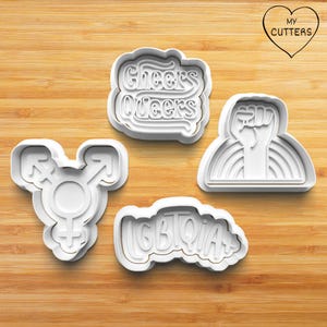 May include: Four white plastic cookie cutters with different designs. The cutters are on a wooden surface. The designs are "Cheers Queers", "LGBTQIA+", a transgender symbol, and a raised fist with a rainbow.