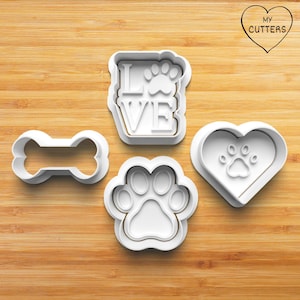 Op de afbeelding: Een set van vier witte plastic koekjesvormpjes met een botvorm, een pootafdruk, een hart met een pootafdruk erin en een rechthoekig frame met de tekst LOVE en een pootafdruk. De tekst MY CUTTERS is zichtbaar in de rechterbovenhoek.