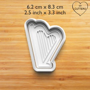 May include: A white harp-shaped cookie cutter with a raised frame and internal string details. The text 6.2 cm x 8.3 cm, 2.5 inch x 3.3 inch, and MY CUTTERS inside a heart shape are visible. The cutter has a clean, moulded plastic construction.