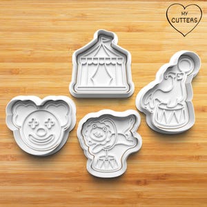 Könnte beinhalten: Vier weiße Ausstecher aus Kunststoff im Zirkus-Thema. Enthält ein Clown-Gesicht, ein Zirkuszelt, einen Löwen auf einer Trommel und einen Seehund, der eine Kugel auf einer Trommel balanciert. Die Ausstecher befinden sich auf einer Holzoberfläche. Der Text "MY CUTTERS" ist herzförmig.