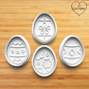 May include: Four white plastic Easter egg-shaped cookie cutters with embossed designs. The designs include a flower, geometric patterns, and wavy lines. The cutters are on a wooden surface. The text "MY CUTTERS" is in a heart-shaped logo.
