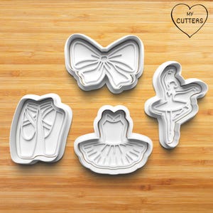 May include: Four white plastic cookie cutters shaped like a ballerina, a ballet shoe, a tutu, and a bow. The cutters are on a wooden surface.