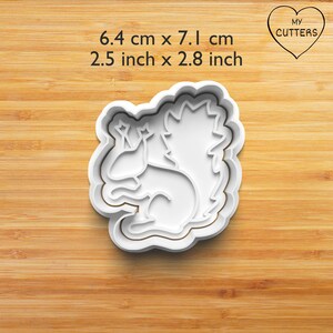 May include: White plastic cookie cutter shaped like a squirrel, measuring 6.4 cm by 7.1 cm. The cutter is on a wooden surface.