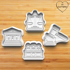 Könnte beinhalten: Vier weiße Ausstecher aus Kunststoff auf einer Holzoberfläche. Die Ausstecher sind in Form eines Gerichtsgebäudes, einer Waage der Gerechtigkeit, eines Richthammer und einer Richterbank mit drei Figuren gehalten. In der oberen rechten Ecke befindet sich ein Herz mit dem Text "MY CUTTERS".