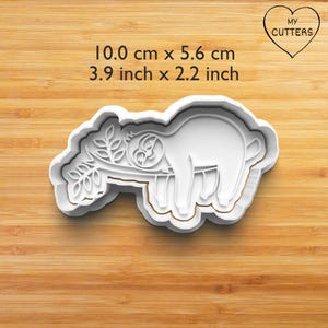 Può includere: Un tagliabiscotti in plastica bianca a forma di bradipo appeso a un ramo. Il testo MY CUTTERS è inserito all'interno di un cuore. Le dimensioni sono 10,0 cm per 5,6 cm. Lo stampo presenta un bordo rialzato per tagliare la pasta.