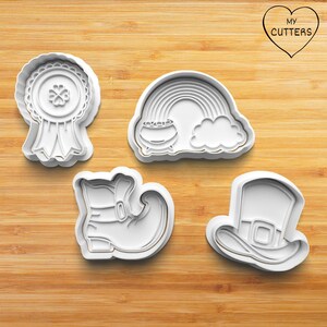 May include: Set of five white plastic cookie cutters shaped like a leprechaun's boot, a pot of gold at the end of a rainbow, a leprechaun's hat, a shamrock with a ribbon, and a rainbow. The cutters are on a wooden surface. My Cutters is printed on a heart-shaped sticker.