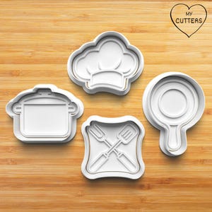May include: Set of four white plastic cookie cutters shaped like a chef's hat, a pot, a frying pan, and a fork and spatula crossed. The cutters are on a wooden surface.