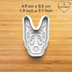 May include: A white Doberman dog head cookie cutter with detailed facial features including pointed ears and a snout. The dimensions are 4.9 cm by 8.0 cm. The text MY CUTTERS is visible inside a heart shape in the upper right corner.