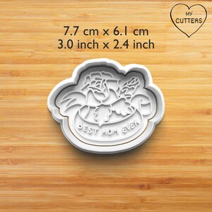 Könnte beinhalten: Ein weißer Ausstechform aus Kunststoff mit einem Rosenmotiv und dem eingeprägten Text "BEST MOM EVER" in der Mitte. Die Form misst 7,7 cm mal 6,1 cm. Das Design zeigt ein detailliertes Blumenmuster mit Blättern und Blütenblättern.