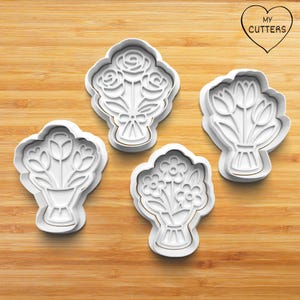 May include: Four white plastic cookie cutters shaped like bouquets of flowers. The cutters are on a wooden surface.