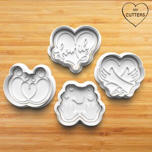 May include: Four white cookie cutters with gold trim are arranged on a wooden surface. The cutters include a bear with a heart, a heart with the word "family", a heart with hugging arms, and a design with hands.