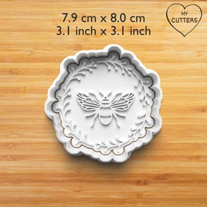 Könnte beinhalten: Weißer Ausstecher in Bienenform, 7,9 cm x 8,0 cm groß, mit dekorativem floralem Rand. Der Ausstecher zeigt ein detailliertes Bienendesign in der Mitte und den Text "MY CUTTERS" in Herzform.