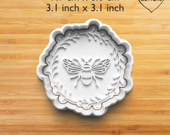 Może przedstawiać: Biała wykrawaczka do ciastek w kształcie pszczoły, o wymiarach 7,9 cm x 8,0 cm, z dekoracyjną kwiatową obwódką. Wykrawaczka posiada szczegółowy wzór pszczoły pośrodku i napis "MY CUTTERS" w kształcie serca.