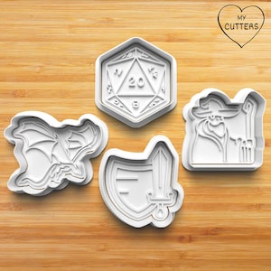 Könnte beinhalten: Vier weiße Ausstecher aus Kunststoff in verschiedenen Formen: ein Drache, ein zwanzigseitiger Würfel, ein Schild und ein Schwert sowie ein Zauberer. Die Ausstecher sind auf einer Holzoberfläche angeordnet. Der Text "MY CUTTERS" befindet sich in einem Herz.