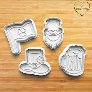 May include: Four white plastic cookie cutters shaped like a leprechaun, a beer mug, a hat, and a shamrock flag. The cutters are on a wooden surface.