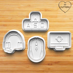 Könnte beinhalten: Vier weiße Plastik-Ausstecher in Form eines Gaming-Headsets, einer Computermaus, eines Computermonitors mit einem Ziel in der Mitte und einer Tastatur mit den Buchstaben "WASD" auf den Tasten.